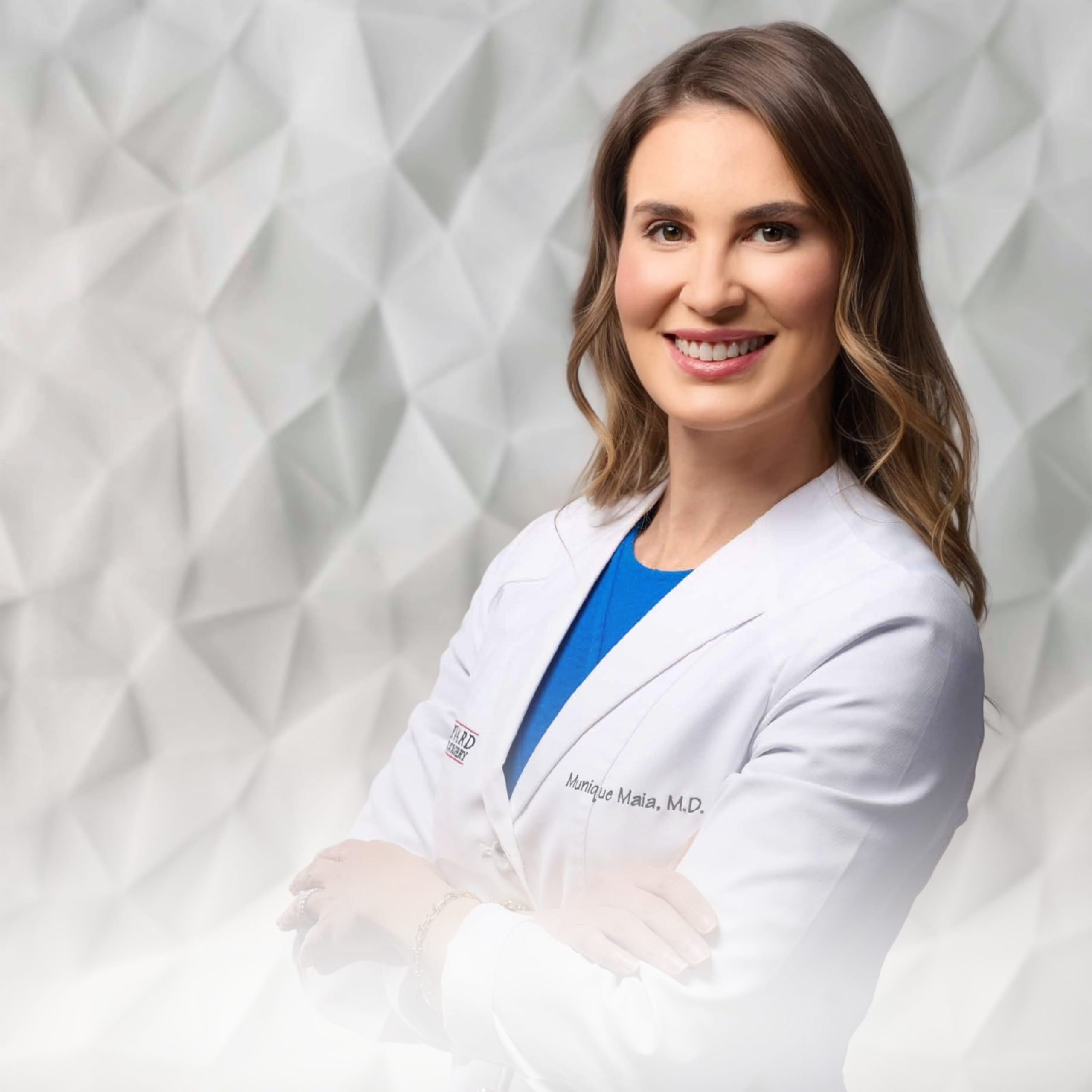 A woman in a white medical coat stands smiling with arms crossed against a geometric-patterned background.