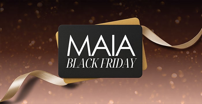 A black card with "MAIA Black Friday" text sits in front of a gold card and ribbon, set against a brown gradient background with sparkles.