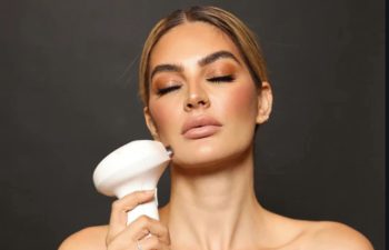 Person holding a white skincare device to their face, eyes closed, with a neutral background.