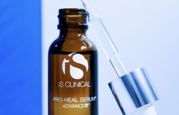 A brown bottle labeled "iS Clinical Pro-Heal Serum Advance+" with a silver dropper cap rests on a white surface against a blue background.